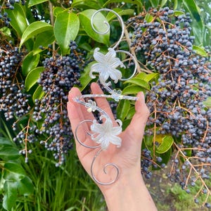 May include: A silver-toned metal arm cuff featuring a spiraling wire design adorned with two large, translucent white floral accents and smaller clear crystal-like embellishments. The structure is flexible and curves to fit around the upper arm.