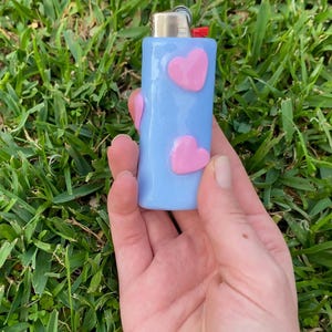 Pode incluir: Um isqueiro retangular azul-claro com dois adornos cor-de-rosa em forma de coração em relevo na parte frontal. O isqueiro possui um mecanismo de ignição superior em metal prateado com uma pequena alavanca vermelha como ferramenta funcional.