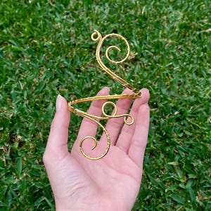 May include: A gold-coloured wire arm cuff featuring a series of interconnected, spiralling scroll designs. The structure is crafted from a single continuous piece of metal wire, wrapped with thinner wire to create a textured, reinforced appearance.