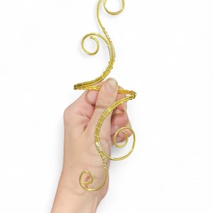 May include: A gold-coloured, wire-sculpted decorative object with a swirling, abstract design. The piece features a central horizontal element and a vertical, curved section with spiral details. The wire is wrapped in a lighter colour in some areas.
