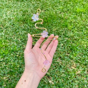 May include: A gold-toned wire arm cuff featuring a spiralling design adorned with three translucent pink flower-shaped charms and several small, multi-coloured crystal beads spaced along the metallic band for a decorative appearance.
