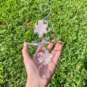 May include: A silver-toned metal arm cuff featuring a spiraling wire design adorned with two translucent, pale pink floral accents. Each flower has a small, iridescent center detail, and the metal wire forms decorative swirls at both ends.