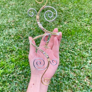 May include: A handcrafted wire sculpture with gold and silver swirls and a star charm. The sculpture features a spiral design with clear crystal accents. The sculpture is held against a backdrop of green grass.
