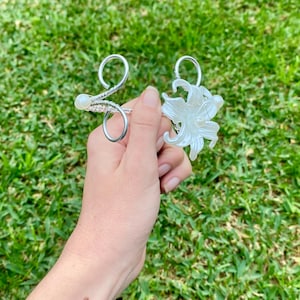 May include: Two silver-toned hair accessories. One has a swirling design with a pearl accent, and the other has a white flower shape with pearl details. The accessories are held against a backdrop of green grass.