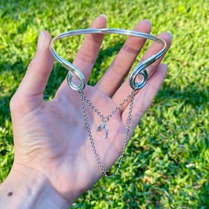 May include: A silver-toned metal choker necklace featuring a rigid circular band with coiled wire accents at the ends. Two delicate silver chains drape from the coils, supporting a single clear, teardrop-shaped crystal pendant at the centre.