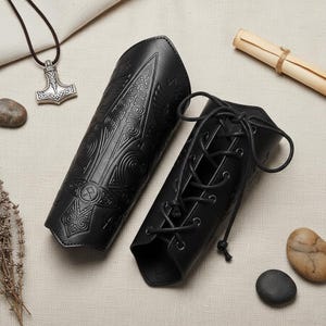 May include: Black leather arm guards with embossed Viking patterns. One is laced, the other a solid sleeve. A silver Thor's hammer pendant and rolled parchment are in the background. The bracers are approximately 25 cm long.
