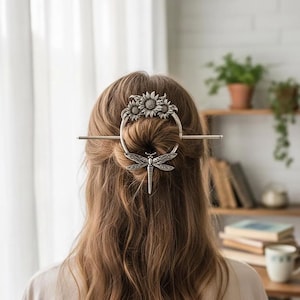 Può includere: Ferro per capelli argentato con un design a libellula e girasole. L'accessorio per capelli è indossato in uno chignon, con il ciondolo a libellula appeso sotto. La parte superiore del ferro per capelli ha un anello con tre girasoli.