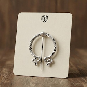 May include: A silver-toned circular penannular brooch featuring an intricate engraved pattern along the ring and curled terminals. The brooch is attached to a light beige display card with a small black shield logo at the top center.