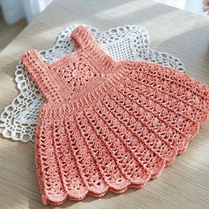 May include: A peach-coloured, handmade crochet dress with a square neckline and a scalloped hem. The dress is placed on a white, lacy doily. The crochet work features a floral design on the bodice and an openwork pattern on the skirt.