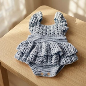 May include: A light blue crochet romper with ruffled detailing. The handmade baby garment features a round neckline, flutter sleeves, and a tiered skirt. The romper has two wooden buttons at the bottom.
