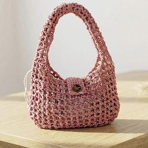 May include: A dusty pink handbag crafted from a woven macrame cord material. The bag features a single integrated handle, a structured base, and a central flap closure secured with a single dark brown button. A light-colored fabric lining is visible.