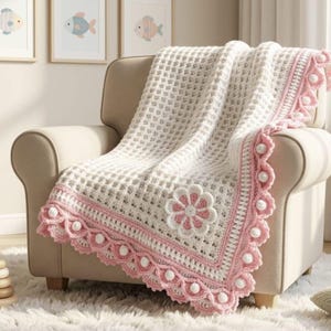 Op de afbeelding: Een witte gehaakte wafelpatroon deken met een roze geschulpte rand met witte pompons en een grote centrale bloemapplicatie in roze en wit. De deken ligt over een beige fauteuil en toont een gestructureerd rasterpatroon en gedetailleerd haakwerk.
