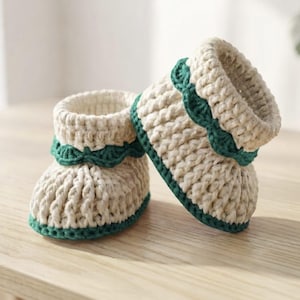 Könnte beinhalten: Ein Paar cremefarbene gehäkelte Babyschuhe mit einem dicken, strukturierten Strickmuster. Jeder Schuh ist mit einer dunkelgrünen Häkelborte am Knöchelbündchen und einem passenden dunkelgrünen Rand an der Sohlenbasis verziert.