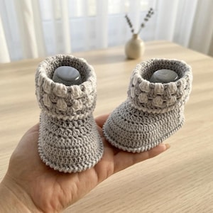 May include: A pair of light grey, hand-crocheted baby booties. The booties feature a textured cuff and a simple design. The soft, neutral colour makes them suitable for any baby.