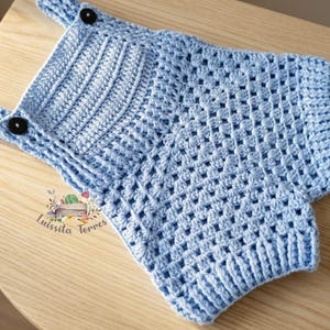Può includere: Un pagliaccetto all'uncinetto azzurro con bottoni neri sulle spalline. L'indumento per bambini fatto a mano è realizzato in filato morbido e presenta un motivo a punto strutturato. Il pagliaccetto è esposto su una superficie di legno chiaro.