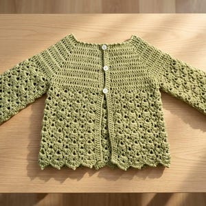 Peut inclure: Un gilet crocheté vert sauge doté de manches longues avec un motif en point coquille texturé, une encolure ronde et une ouverture verticale sur le devant fermée par trois petits boutons blancs. Le vêtement présente une maille uniforme.