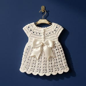 Può includere: Un vestito da neonato bianco all'uncinetto con un grande fiocco di raso e abbellimenti decorativi. L'abito è appeso a un gancio dorato su uno sfondo blu scuro. L'abito ha un orlo smerlato.
