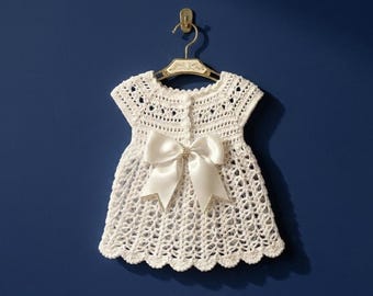 May include: A white, crocheted baby dress with a large satin bow and decorative embellishments. The dress is hanging on a gold hanger against a dark blue background. The dress has a scalloped hemline.
