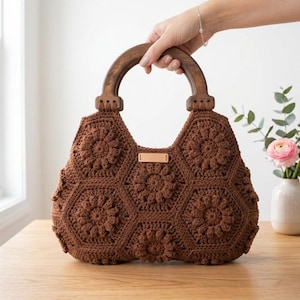 May include: A brown crochet handbag constructed from multiple hexagonal floral motifs. The bag features a dark brown, semi-circular wooden handle attached to the top. A small, rectangular, light-colored leather tag is centered on the front panel.