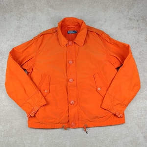 May include: Bright orange jacket with a button-up front, two front pockets, and a collared neckline. The jacket has button details on the cuffs and shoulders. A drawstring is visible at the bottom hem.
