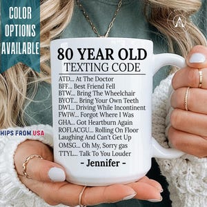 May include: A white ceramic mug featuring black text that reads 80 YEAR OLD TEXTING CODE followed by a list of humorous acronyms and definitions, ending with the name Jennifer. The mug has a standard handle and a smooth, glossy finish.
