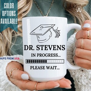 May include: A white ceramic mug featuring a black line drawing of a graduation cap at the top. Below the cap, the text reads DR. STEVENS IN PROGRESS... followed by a loading bar graphic and the text PLEASE WAIT... in bold black lettering.