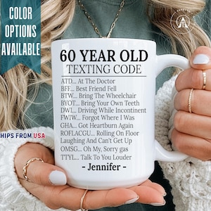 May include: A white ceramic mug featuring black printed text titled 60 YEAR OLD TEXTING CODE, followed by a list of humorous acronyms and their meanings, including ATD, BFF, BTW, BYOT, DWI, FWIW, GHA, ROFLACGU, OMSG, and TTYL, ending with Jennifer.