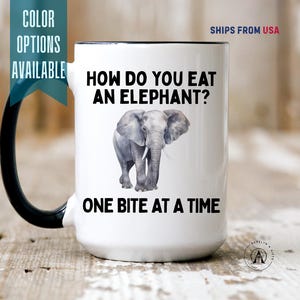 May include: A white ceramic mug with a black handle and interior. The front features a gray elephant illustration centered between the black text "HOW DO YOU EAT AN ELEPHANT?" at the top and "ONE BITE AT A TIME" at the bottom in a bold sans-serif font.