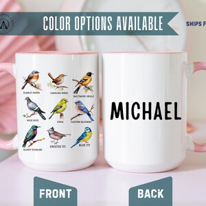 May include: Two white ceramic mugs with light pink handles and rims. The front mug features a grid of nine colorful bird illustrations with labels including Scarlet Robin, Carolina Wren, Baltimore Oriole, Rock Dove, Finch, Eastern Bluebird, Glossy Starling, Crested Tit, and Blue Tit. The back mug displays the name MICHAEL in bold black capital letters.
