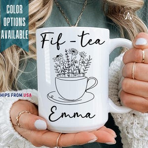 May include: White ceramic mug with the text "Fif-tea Emma" and a floral design inside a teacup. The mug has a large handle and is held by a person. The words "Color Options Available" are visible on the left side of the mug.