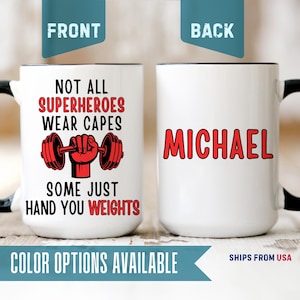 May include: A white ceramic mug with a black handle and rim. The front features a red graphic of a hand lifting a barbell with the black and red text: NOT ALL SUPERHEROES WEAR CAPES SOME JUST HAND YOU WEIGHTS. The back displays the name MICHAEL in red.