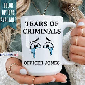 May include: A white ceramic mug featuring the black text TEARS OF CRIMINALS at the top and OFFICER JONES at the bottom. Between the text lines is a graphic of two cartoon eyes with blue tears streaming down from them on a white background.