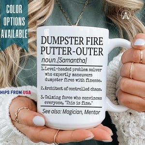 May include: White ceramic mug with the text "DUMPSTER FIRE PUTTER-OUTER" and a definition. The mug also includes the name "Samantha" and a list of characteristics. The mug is held by a person.