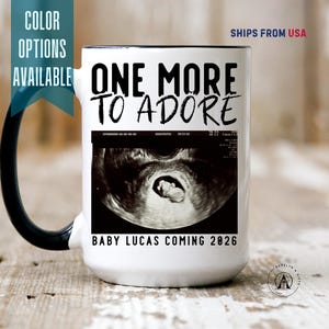 May include: White ceramic mug with a black handle and rim. The mug features the text "ONE MORE TO ADORE" above an ultrasound image. Below the image, it reads "BABY LUCAS COMING 2026". Color options are available.