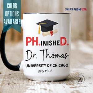 May include: A white ceramic mug with a black handle featuring a graphic of a black graduation cap and a diploma. The text reads PH.INISHED. Dr. Thomas, UNIVERSITY OF CHICAGO, and Est. 2026 in black and red lettering on the front of the mug.