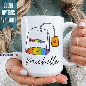 May include: A white ceramic mug featuring a graphic of a tea bag with a rainbow-colored bottom and a small smiling face. The text "LGBTea" is printed in rainbow letters on the tea bag, with the name "Michelle" written in black script below.