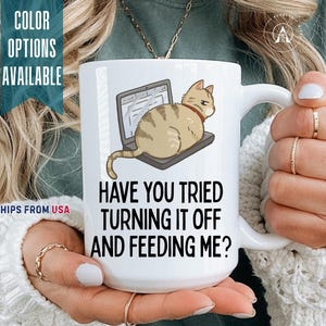 May include: A white ceramic mug featuring a graphic of a tan tabby cat sitting on a laptop. Below the cat, the text reads: HAVE YOU TRIED TURNING IT OFF AND FEEDING ME? The mug has a curved handle and a smooth, glossy finish with black lettering.