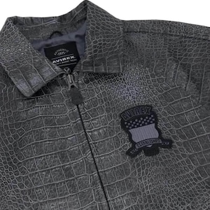 Puede incluir: Chaqueta gris de piel sint&eacute;tica con textura de piel de cocodrilo. La chaqueta tiene cierre de cremallera y cuello. Un parche en el pecho dice "AVIREX ESTABLISHED 1975".