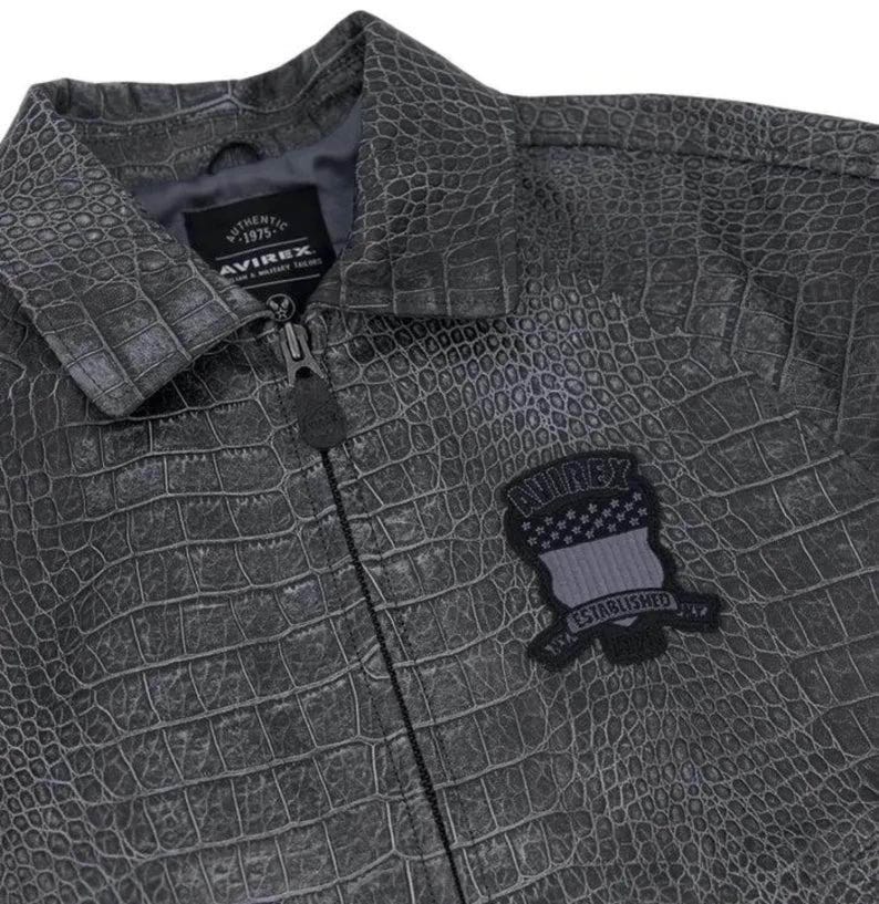Puede incluir: Chaqueta gris de piel sint&eacute;tica con textura de piel de cocodrilo. La chaqueta tiene cierre de cremallera y cuello. Un parche en el pecho dice "AVIREX ESTABLISHED 1975".