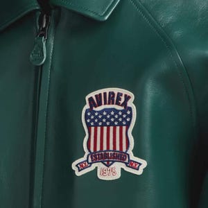 May include: A close-up of a dark green leather jacket with a collar and a zip. The jacket has a patch with the text "AVIREX ESTABLISHED NY 1975" in red, white, and blue.