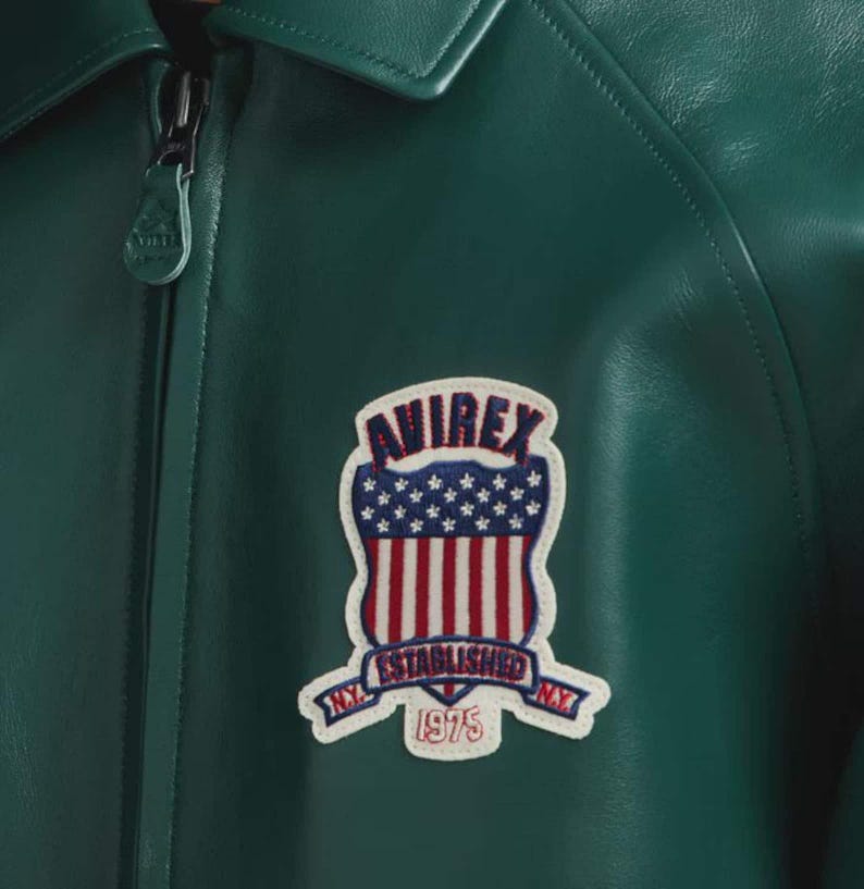 May include: A close-up of a dark green leather jacket with a collar and a zip. The jacket has a patch with the text "AVIREX ESTABLISHED NY 1975" in red, white, and blue.