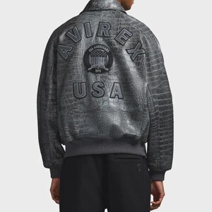 Puede incluir: Chaqueta bomber gris con textura de piel de cocodrilo. La parte trasera de la chaqueta presenta el texto "AVIREX" arqueado sobre un emblema circular y la palabra "USA". La chaqueta tiene dobladillo y pu&ntilde;os de canal&eacute;.