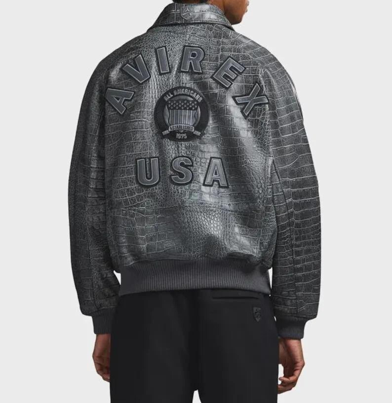 Puede incluir: Chaqueta bomber gris con textura de piel de cocodrilo. La parte trasera de la chaqueta presenta el texto "AVIREX" arqueado sobre un emblema circular y la palabra "USA". La chaqueta tiene dobladillo y pu&ntilde;os de canal&eacute;.