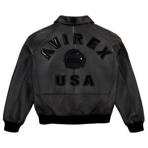 Pu&ograve; includere: Giacca bomber in pelle nera con la scritta "AVIREX" ad arco sulla schiena, sopra un emblema circolare e la scritta "USA". La giacca ha colletto, polsini e orlo a coste.