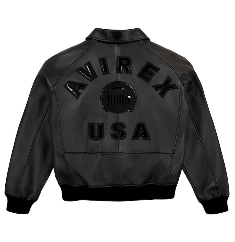 Pu&ograve; includere: Giacca bomber in pelle nera con la scritta "AVIREX" ad arco sulla schiena, sopra un emblema circolare e la scritta "USA". La giacca ha colletto, polsini e orlo a coste.