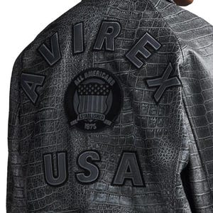 May include: A dark gray jacket with a crocodile skin texture. The back of the jacket features the words "AIR," "REX," and "USA" in large, raised letters. A circular patch with the words "ALL AMERICANS" and "ESTABLISHED 1975" is also visible.