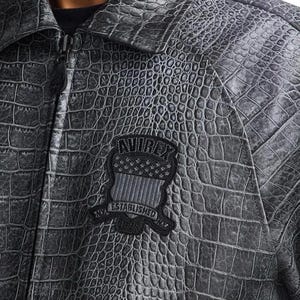 Puede incluir: Chaqueta gris de imitaci&oacute;n de cocodrilo con cierre de cremallera y cuello. La chaqueta presenta un parche con el texto "AVIREX ESTABLISHED NYC 1975" en negro y gris.