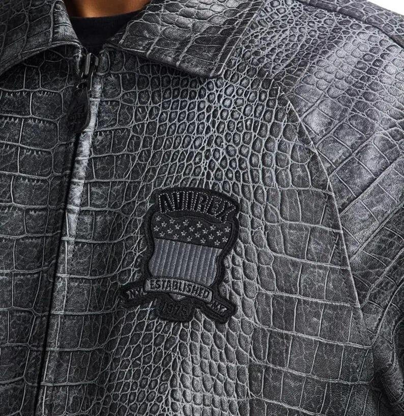 Puede incluir: Chaqueta gris de imitaci&oacute;n de cocodrilo con cierre de cremallera y cuello. La chaqueta presenta un parche con el texto "AVIREX ESTABLISHED NYC 1975" en negro y gris.