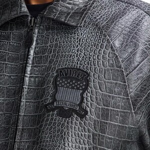 May include: A gray, textured jacket with a crocodile skin pattern. The jacket has a zipper closure and a collar. A black patch with the text "AVIREX ESTABLISHED 1975" and a flag design is sewn on the chest. The jacket is made of a durable material.