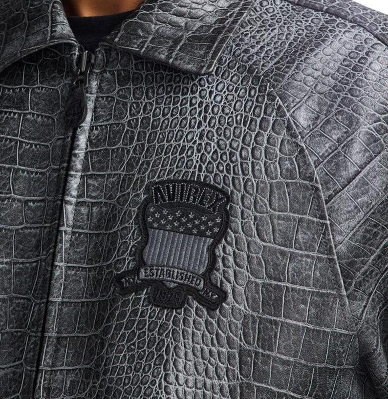 May include: A gray, textured jacket with a crocodile skin pattern. The jacket has a zipper closure and a collar. A black patch with the text "AVIREX ESTABLISHED 1975" and a flag design is sewn on the chest. The jacket is made of a durable material.